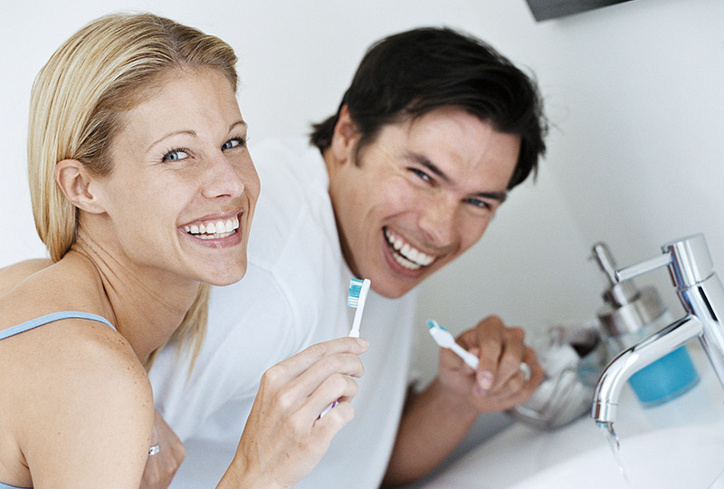 Two people brushing their teeth in a bathroom with a faucet and toothbrushes.