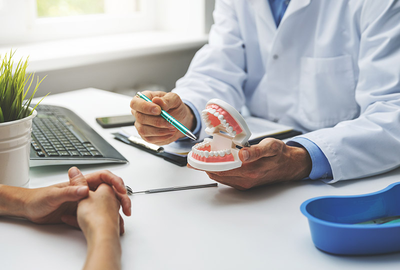The image shows a dental professional examining a patient s teeth with a dental mirror while seated at a desk in an office setting, with a person sitting across from them and another individual observing the examination.